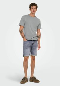 Camiseta de algodón gris con cuello redondo, combinada con pantalones cortos de mezclilla gris claro con dobladillos doblados. Unos zapatos de ante marrón completan el atuendo.