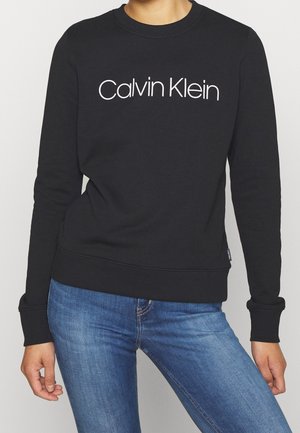 Czarna bluza z długim rękawem z logo "Calvin Klein" w białym kolorze na wysokości klatki piersiowej, noszona z niebieskimi dżinsami przez stojącą osobę.