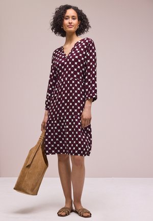 Vrouw met krullend haar die een knielange bordeauxrode jurk met wit patroon draagt, een lichtbruine suède tas vasthoudend, staand tegen een neutrale achtergrond.
