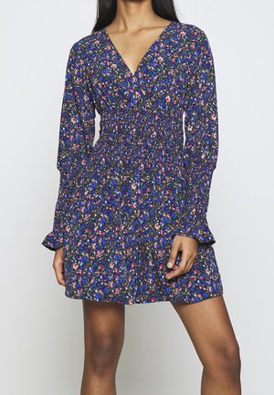 Mujer con vestido corto de manga larga y escote en V con estampado floral en azul, rosa y negro y cintura fruncida sobre un fondo liso.