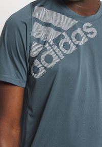 Man som bär en blå t-shirt med kort ärm från Adidas, med en stor vit Adidas-logotyp över bröstet och axeln.
