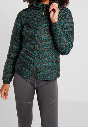 Personne portant une veste matelassée verte et noire à imprimé léopard avec une fermeture éclair à l'avant, associée à des leggings ajustés gris foncé.