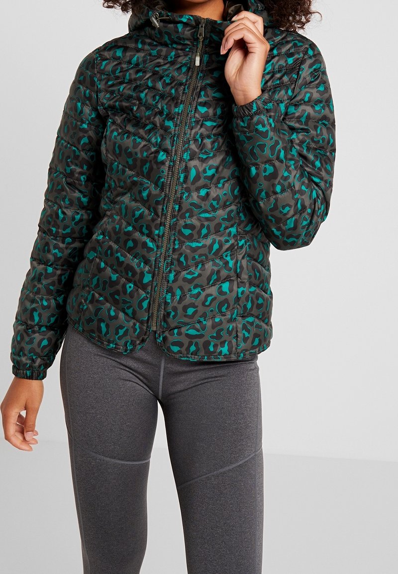 Personne portant une veste matelassée verte et noire à imprimé léopard avec une fermeture éclair à l'avant, associée à des leggings ajustés gris foncé.