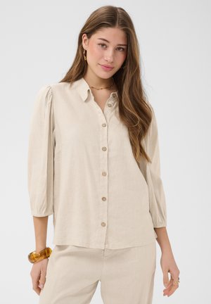 Junge Frau mit langen braunen Haaren trägt eine beige Bluse mit Knöpfen und passende Hose, ergänzt durch ein Bernsteinarmband und goldene Creolen.