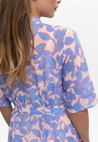 Robe portefeuille florale en tissu transparent avec des feuilles bleues et roses sur un fond clair, dotée de manches courtes et d'une taille ceinturée.