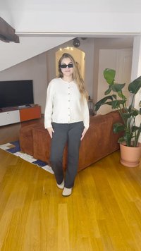 Gaiši bēšs kardigans ar pogām, melnas bikses un krēmkrāsas apavi. Fonā ir brūns dīvāns, telpaugi un televizors.