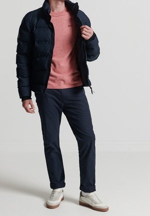 Homme portant une doudoune noire par-dessus un pull corail, un pantalon bleu marine et des baskets blanches, debout devant un fond clair uni.