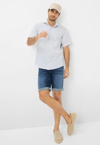 Chemise à manches courtes, rayée bleu clair, shorts en denim avec des ourlets retroussés et chaussures beiges à enfiler. Une casquette neutre complète l'ensemble.