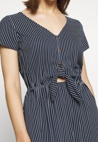 Mörkblå klänning med vita vertikala ränder, V-ringning, korta ärmar, framknäppning och ett knyt i midjan. Slät tygtextur.
