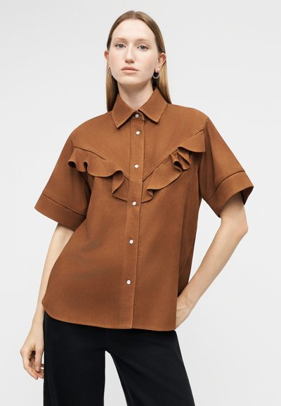 Mujer con camisa marrón de manga corta y botones, con detalles de volantes en el pecho, combinada con pantalones negros, sobre un fondo blanco.