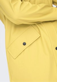 Gul vandtæt jakke med en glat tekstur, der har en stor klaplomme sikret med en sort knap og et strømlinet design.