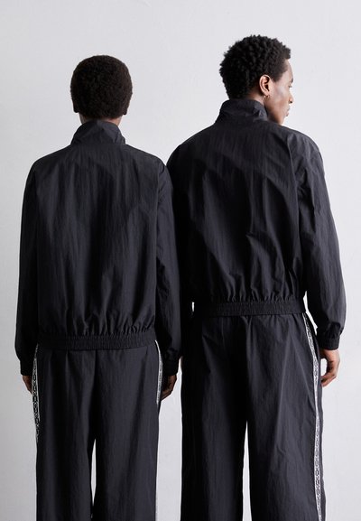 Deux individus portant des survêtements noirs vus de dos, avec des poignets et des ceintures élastiques, et des détails latéraux blancs sur le pantalon.