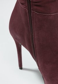 Bota de tacón alto de ante burdeos con cremallera lateral y textura suave. Presenta punta afilada y un diseño sin costuras para una apariencia elegante.