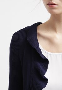 Námořnicky modrý žebrovaný kardigan s detailem volánového límce, nošený přes bílý top. Zaměření na strukturovanou látku a designové prvky.