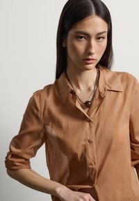 Joven mujer con cabello largo y oscuro, que lleva una camisa marrón abotonada y un collar de cordón negro con un colgante redondo de madera.