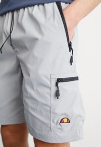 Shorts gris clair en tissu léger, avec deux poches zippées, une taille à cordon de serrage et un logo brodé décoratif en orange et jaune.