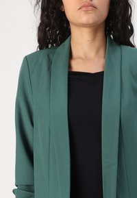 Blazer verde in un tessuto liscio con una vestibilità strutturata e colletto a scialle. Indossato sopra un top nero, caratterizzato da linee semplici e senza chiusure visibili.