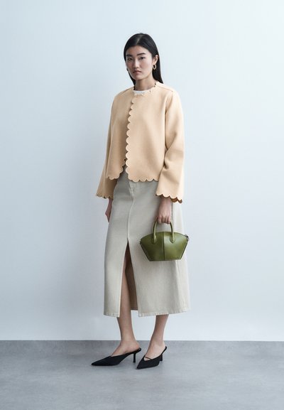 Γυναίκα με μπεζ σακάκι και φούστα με κυματιστό τελείωμα και μπροστινή σχισμή, κρατώντας μικρή πράσινη τσάντα, στέκεται μπροστά σε ένα απλό ανοιχτόχρωμο τοίχο.