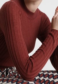 Rode ribgebreide trui met een hoge halslijn, voorzien van lange mouwen. Gecombineerd met een gestreepte broek met cirkelvormige patronen in rood en blauw.