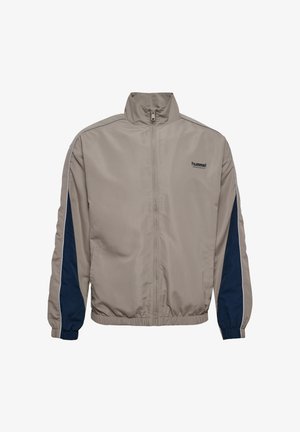 Beżowa kurtka zapinana na zamek z wysokim kołnierzem, granatowymi panelami po bokach, elastycznymi mankietami i dołem, z małym logo na piersi. Gładka faktura tkaniny.