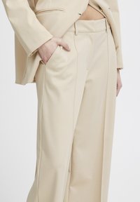 Beige tailored pants made of soft fabric, featuring a smooth texture, front pleats, and side pockets, paired with a matching blazer.