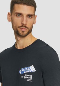 Camiseta de algodón azul marino con un estampado gráfico en blanco y azul que dice "ARTS & SERVICE AND SUPPLIES" en el área del pecho izquierdo. Mangas cortas.
