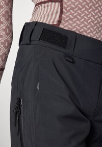 Black outdoor pants with a textured waistband featuring a velcro closure and a zippered pocket on the side. Smooth, water-resistant fabric.