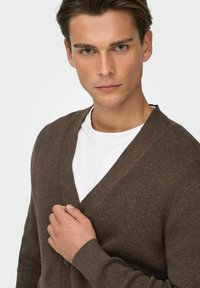 Brun strikket cardigan med V-hals, utfrest med en enkelt knappelukking og ribbestrikkede mansjetter, brukt over en hvit T-skjorte.