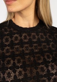 Vrouw met blond haar die een zwarte bloemenkanttop draagt, waarbij ze haar decolleté en een deel van haar gezicht laat zien, licht naar links gekeerd.