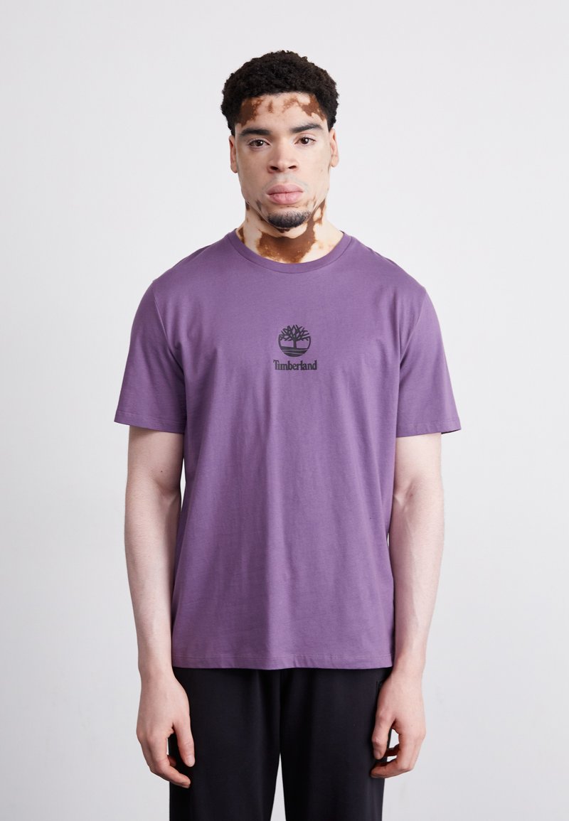 Violet medvilnės marškinėliai su apvalia iškirpte, viduryje krūtinės yra juodas Timberland logotipas. Modelis vilki juodus kelnes.