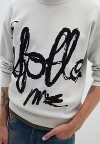 Suéter gris claro con un audaz diseño de pincelada negra que lleva el texto "sígueme". Con puños y dobladillo acanalados, usado sobre jeans azules.