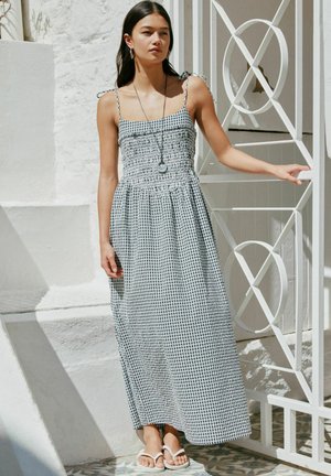 Kobieta w czarno-białej, bezrękawnikowej sukience maxi w kratę gingham i białych klapkach stojąca przy białej metalowej bramie w jasnym świetle słonecznym.