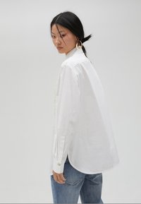 White cotton shirt with long sleeves, relaxed fit, round hem, and button cuffs, paired with blue denim jeans. Simple, classic design.