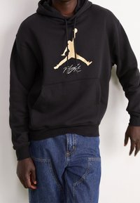 Uomo che indossa un hoodie nero con il logo Jumpman dorato e la scritta "Flight", abbinato a jeans blu, in piedi contro un muro bianco.