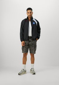 Black windbreaker jacket with white and red accents, gray patterned cargo shorts, and gray sneakers with white soles and socks.