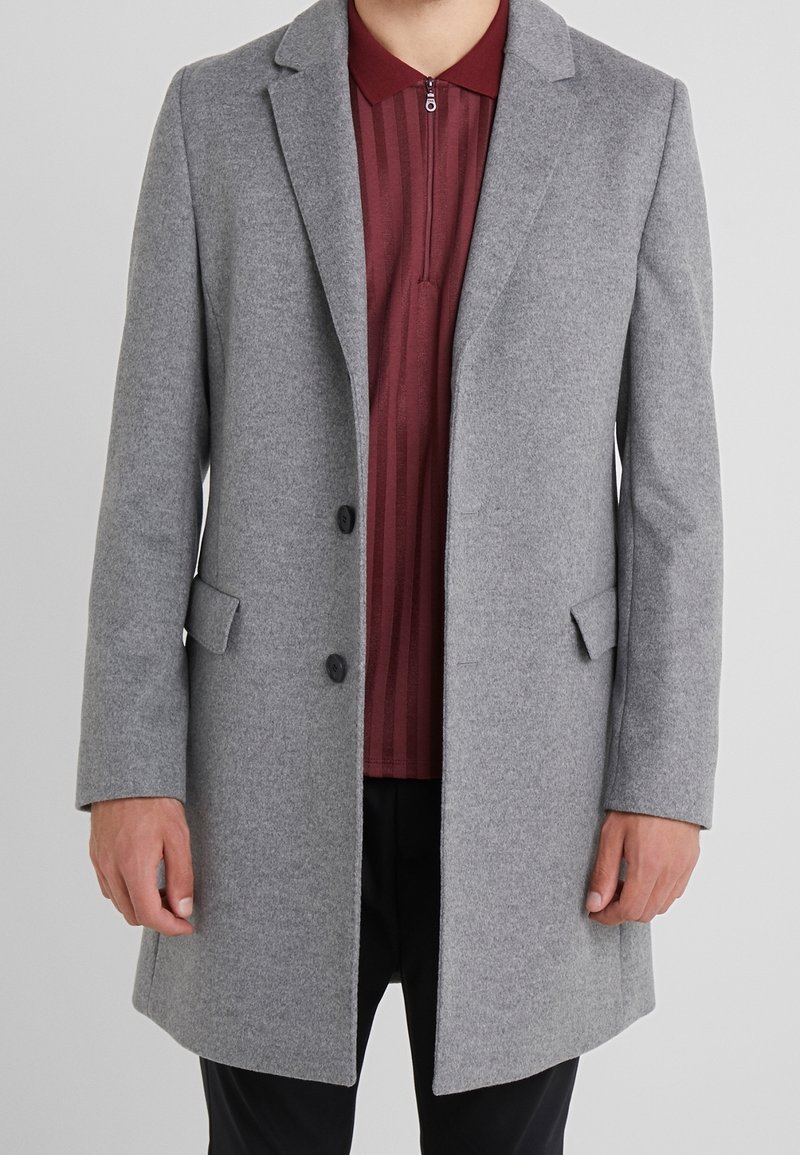 Abrigo de lana gris con solapas muesca, dos botones frontales y bolsillos laterales, llevado sobre una camisa burdeos con cuello con cremallera. Textura suave.