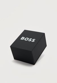 Μαύρο κυβοειδές κουτί με τη λέξη «BOSS» σε έντονα λευκά γράμματα στην κορυφή, τοποθετημένο σε απλό φωτεινό φόντο.