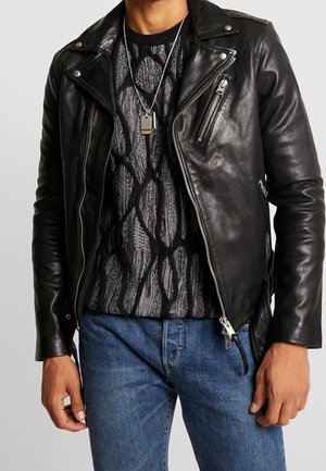 Man met een zwart leren bikerjack over een zwart-grijze gemêleerde trui, blauwe spijkerbroek en een zilveren dogtag ketting.