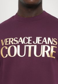 Felpa marrone con collo a girocollo a costine e grande scritta dorata "VERSACE JEANS COUTURE" sul davanti. Tessuto morbido.