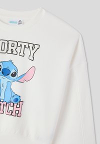 Maglia bianca con un personaggio cartone animato blu con orecchie grandi, scritte sovradimensionate che riportano "SPORTY BITCH" e colletto e polsini a costine.