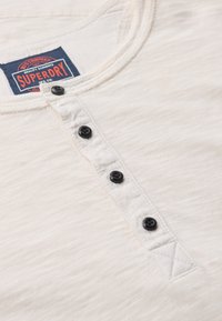 Korte mouwen crèmekleurig shirt met een ronde hals, zwarte knopen, textuurstof en een 'Superdry' merklabel.