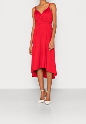 Femme portant une robe portefeuille rouge sans manches avec un ourlet asymétrique et des talons blancs à bride cheville, debout sur un sol gris.