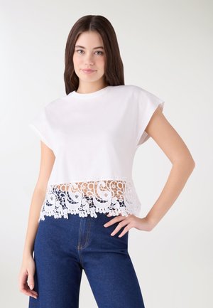 Giovane donna con indosso un top bianco senza maniche con orlo in pizzo dettagliato e jeans blu scuro, in piedi con una mano sul fianco.