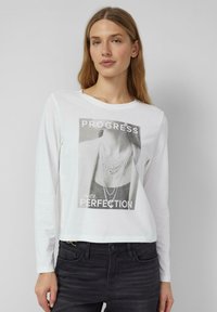 Biała koszulka o długim rękawie z czarno-białym nadrukiem graficznym z napisem "PROGRES ponad DOSKONAŁOŚĆ" i wzorem naszyjnika.