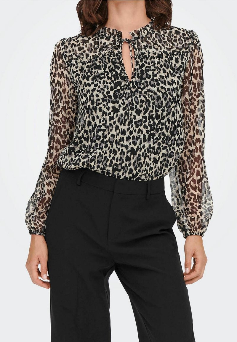 Blouse transparente à manches longues avec un imprimé léopard beige et noir, dotée d'un détail de nœud en forme de trou de serrure et de poignets élastiques. Associée à un pantalon noir.