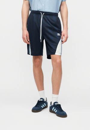 Muž nosiaci námornícke modré športové šortky s bielymi bočnými panelmi, bielou šnúrkou, bielymi ponožkami a námorníckymi modrými teniskami s svetlomodrými pruhmi.