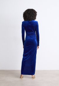 Vestido longo e justo em veludo azul royal, com mangas longas e detalhes franzidos na cintura, apresentando uma textura suave e um design até ao chão.