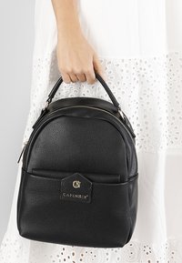 Svart läderryggsäck med en texturerad yta, rundad topp, dubbla handtag och en logotypaccent framtill. Utrustad med guldfärgad hårdvara och dragkedja.