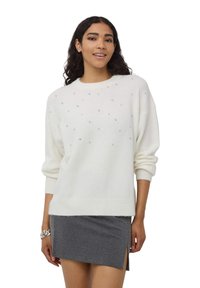 Biała sweterka z dzianiny z rozproszonymi małymi srebrnymi ozdobami, okrągłym dekoltem i obniżoną linią ramion, zestawiona z szarym mini spódnicą.
