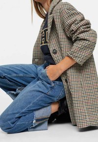 Ruudullinen villakangastakki suurilla napit, tumman hupparin päällä. Siniset kuluneet farkut vetoketjuyksityiskohdalla ja käärityillä lahkeilla. Mustat saappaat näkyvissä.